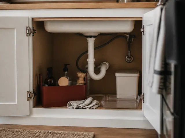 An open under-sink cabinet with visible plumbing, a burnt-sienna caddy, cloths, a tray, and a small bin